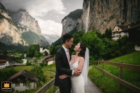 In Interlaken, Switzerland, a couple poses together on a path framed by rustic wooden railings, with a charming Swiss village and a picturesque waterfall cascading in the background.